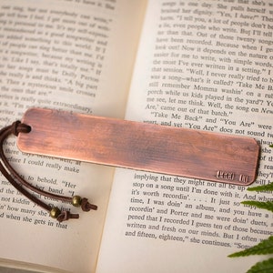 May include: A copper bookmark with a brown leather strap and brass beads. The bookmark has the text "4444 00" engraved on it.
