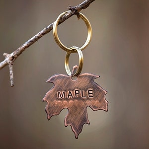 May include: A copper maple leaf keychain with the word "MAPLE" engraved on it. The keychain is hanging from a branch.