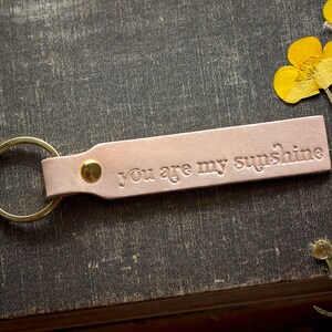 May include: A light pink leather keychain with a gold ring and the words "You are my sunshine" embossed on the leather.