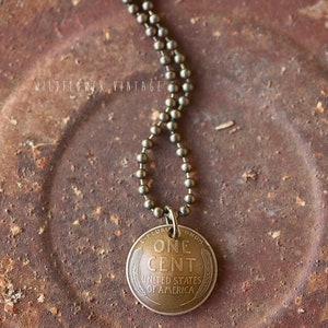 May include: A gold chain necklace with a vintage one cent coin pendant. The coin has the words "One Cent" and "United States of America" inscribed on it.