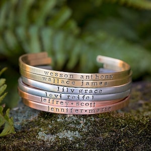 May include: Stack of personalized metal cuff bracelets in gold, silver, and copper tones. Each bracelet is engraved with a name. The bracelets are stacked on a mossy surface with a blurred green background.