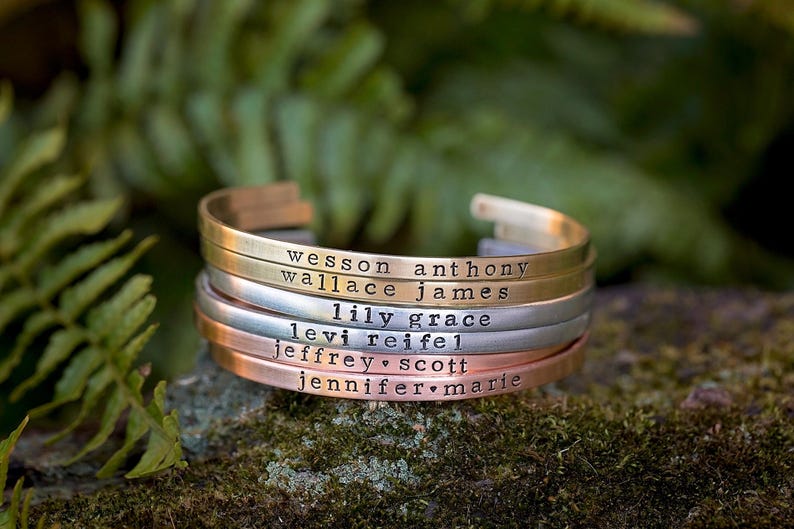 May include: Stack of personalized metal cuff bracelets in gold, silver, and copper tones. Each bracelet is engraved with a name. The bracelets are stacked on a mossy surface with a blurred green background.