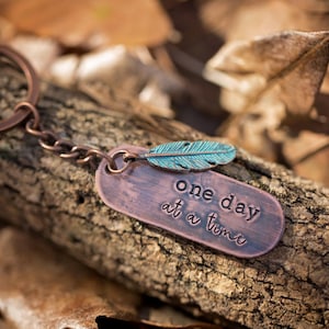 May include: A copper keychain with a feather charm and a tag that reads "one day at a time".
