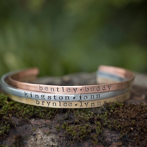 May include: Three personalized cuff bracelets in copper, silver, and gold tones, each engraved with names and a heart symbol. The bracelets are displayed on a mossy, natural surface, with a blurred green background.
