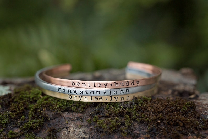 May include: Three personalized cuff bracelets in copper, silver, and gold tones, each engraved with names and a heart symbol. The bracelets are displayed on a mossy, natural surface, with a blurred green background.