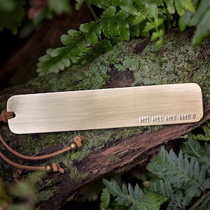 May include: A rectangular brushed brass bookmark with rounded corners featuring a series of etched tally marks on the lower right side. A brown leather cord is threaded through a hole on the left end, secured with two small brass beads.