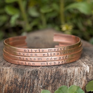 May include: Four copper-colored metal cuff bracelets stacked together. Each bracelet is engraved with a name: Olivia Maeve, Aveen Elizabeth, Graham Edward, and Quinn Michael. The bracelets are displayed on a wooden surface.
