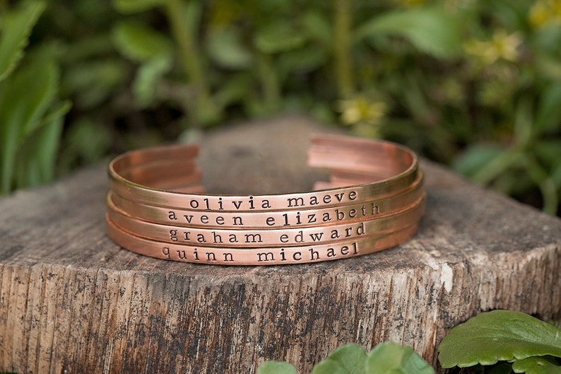 May include: Four copper-colored metal cuff bracelets stacked together. Each bracelet is engraved with a name: Olivia Maeve, Aveen Elizabeth, Graham Edward, and Quinn Michael. The bracelets are displayed on a wooden surface.
