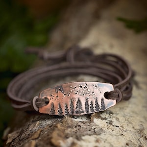 May include: A copper-colored bracelet with a forest and night sky design. The rectangular metal plate features etched trees, stars, and a crescent moon. The bracelet has a brown leather cord that wraps around the wrist.