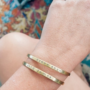 May include: Two gold-colored metal cuff bracelets are worn on a wrist. The top bracelet reads "greer sean" and the bottom bracelet reads "reid james" in a simple, sans-serif font. The bracelets are slightly curved to fit the wrist.