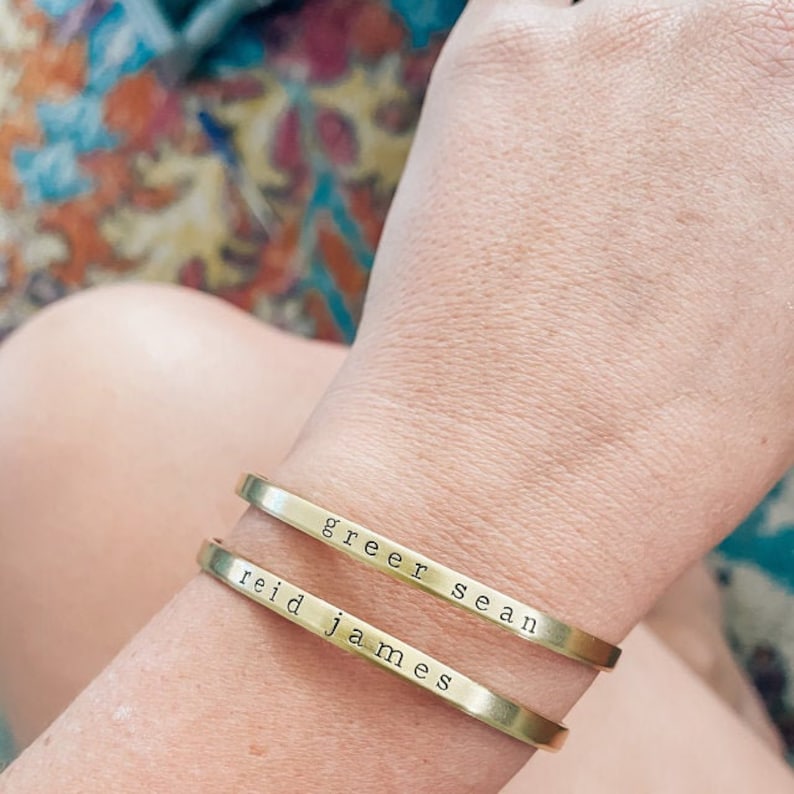 May include: Two gold-colored metal cuff bracelets are worn on a wrist. The top bracelet reads "greer sean" and the bottom bracelet reads "reid james" in a simple, sans-serif font. The bracelets are slightly curved to fit the wrist.