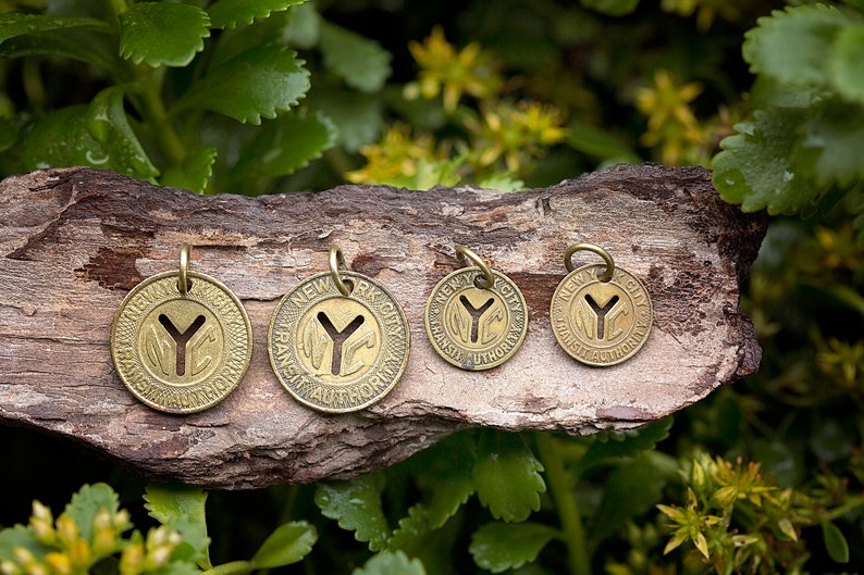 May include: Four brass charms with the New York City Transit Authority logo, featuring a stylized "Y" and the words "New York City Transit Authority". The charms are different sizes and are arranged on a piece of wood.