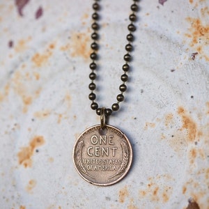 May include: A copper-colored pendant necklace featuring a vintage US one-cent coin with the inscription "ONE CENT UNITED STATES OF AMERICA". The necklace is made with a simple chain.