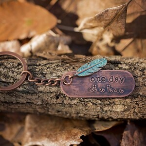 May include: A copper keychain with a turquoise feather charm and a copper tag that reads "one day at a time".