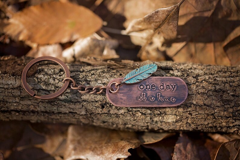 May include: A copper keychain with a turquoise feather charm and a copper tag that reads "one day at a time".