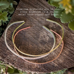 May include: Three aluminum bracelets in silver, gold, and copper tones rest on a wooden surface surrounded by greenery. The bracelets are open-ended cuffs. Text on the image states, "Aluminum bracelets are thicker, since aluminum is a softer metal than copper and brass."