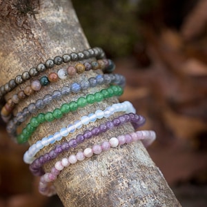 May include: A stack of colorful beaded bracelets made of various gemstones, including amethyst, rose quartz, and green aventurine. The bracelets are arranged on a piece of tree bark.