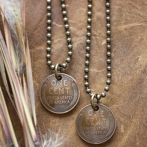 May include: Two gold chain necklaces with a copper penny pendant on each. The pennies are from the United States of America and say "One Cent" on them.