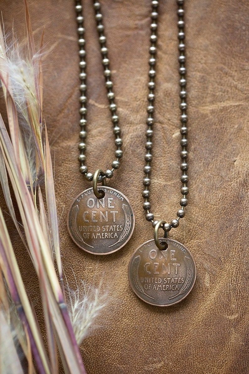 May include: Two gold chain necklaces with a copper penny pendant on each. The pennies are from the United States of America and say "One Cent" on them.