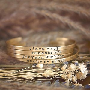 May include: Four gold-colored metal cuff bracelets stacked together. Each bracelet is engraved with a name: clare noel, ransom ark, grayson embrey, and zachary scott. The bracelets are resting on dried flowers and grasses.
