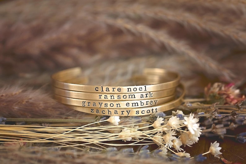 May include: Four gold-colored metal cuff bracelets stacked together. Each bracelet is engraved with a name: clare noel, ransom ark, grayson embrey, and zachary scott. The bracelets are resting on dried flowers and grasses.