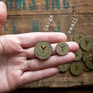 May include: Two brass tokens with the words "Good For One Fare" and "New York City" on them. The larger token is in the left hand, and the smaller token is in the right hand. A pile of smaller tokens is in the background.