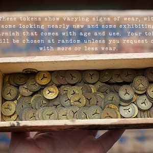 May include: A wooden box filled with a large quantity of brass tokens. The tokens are all circular and have a Y-shaped design in the center. The tokens show varying signs of wear, with some looking nearly new and some exhibiting tarnish that comes with age and use.
