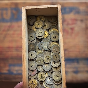 May include: A wooden box filled with a large quantity of old, gold-colored metal tokens. The tokens have a small hole in the center and a design on the front.