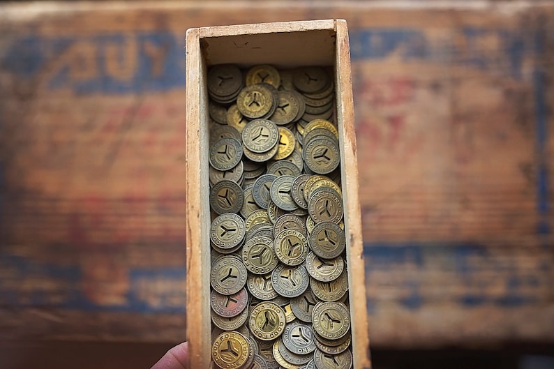 May include: A wooden box filled with a large quantity of old, gold-colored metal tokens. The tokens have a small hole in the center and a design on the front.