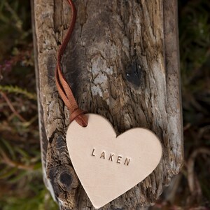 May include: A light brown leather heart-shaped tag with a brown leather cord. The tag has the word "LAKEN" embossed on it.