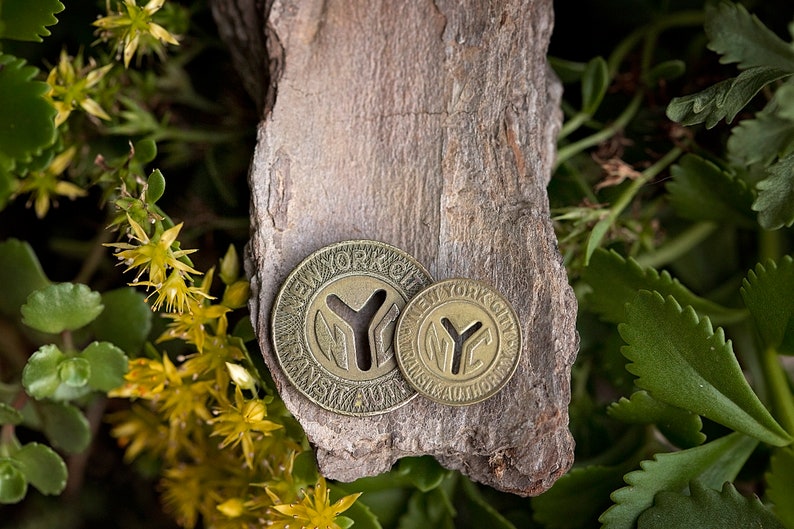 May include: Two gold-colored metal tokens with the words "New York City Transit Authority" and the letter "Y" in a circle. The tokens are on a piece of bark.