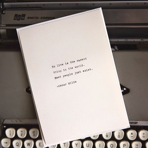 May include: A white card with a quote from Oscar Wilde typed on a vintage typewriter. The quote reads: "To live is the rarest thing in the world. Most people just exist."