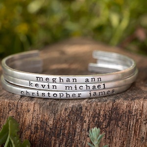 May include: Three silver-colored metal cuff bracelets stacked on a wooden surface. Each bracelet is engraved with a name: Meghan Ann, Kevin Michael, and Christopher James. The background is blurred green foliage.