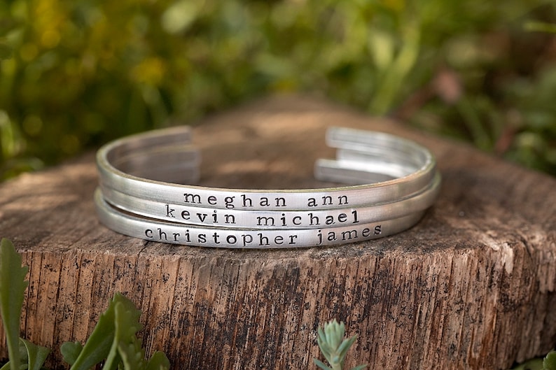 May include: Three silver-colored metal cuff bracelets stacked on a wooden surface. Each bracelet is engraved with a name: Meghan Ann, Kevin Michael, and Christopher James. The background is blurred green foliage.