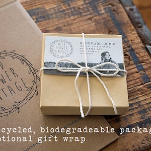 May include: A small, tan gift box tied with white twine, featuring a label with the text "HANDMADE GOODS" and "WILD FLOWER VINTAGE." The box is on a rustic wooden surface, with a larger cardboard box in the background.