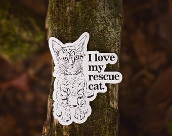 Puede incluir: Una pegatina blanca con una ilustración en blanco y negro de un gato y el texto "Amo a mi gato de rescate."