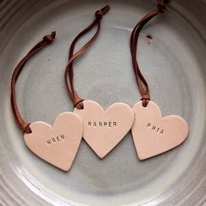May include: Three light brown leather heart-shaped tags with brown leather strings. The tags are engraved with the names "Wren", "Harper", and "Phia".