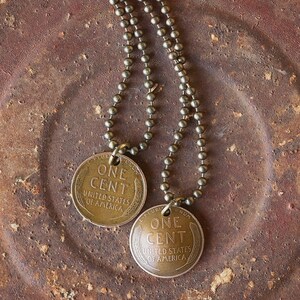 May include: A necklace with two antique-looking copper-colored one-cent coins as pendants. The coins are attached to a chain made of small, round beads. The necklace is lying on a rusty metal surface.