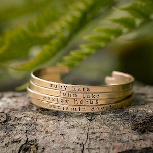 May include: A gold-colored cuff bracelet with four stacked bands, each engraved with a name: Ruby Kate, John Luke, Wesley Hobbs, and Benjamin David. The bracelet rests on a piece of wood, with green foliage in the background.