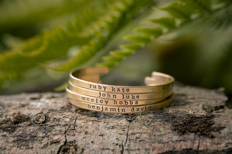 May include: A gold-colored cuff bracelet with four stacked bands, each engraved with a name: Ruby Kate, John Luke, Wesley Hobbs, and Benjamin David. The bracelet rests on a piece of wood, with green foliage in the background.