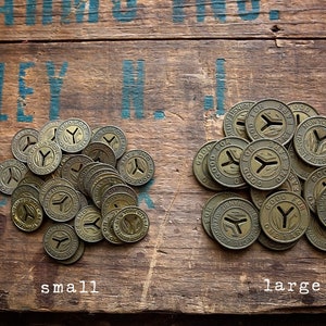 May include: Two piles of vintage brass tokens on a wooden surface. The tokens are round and have a Y-shaped design in the center. The text "GOOD FOR ONE FARE" is visible on the tokens. The pile on the left is labeled "small" and the pile on the right is labeled "large".