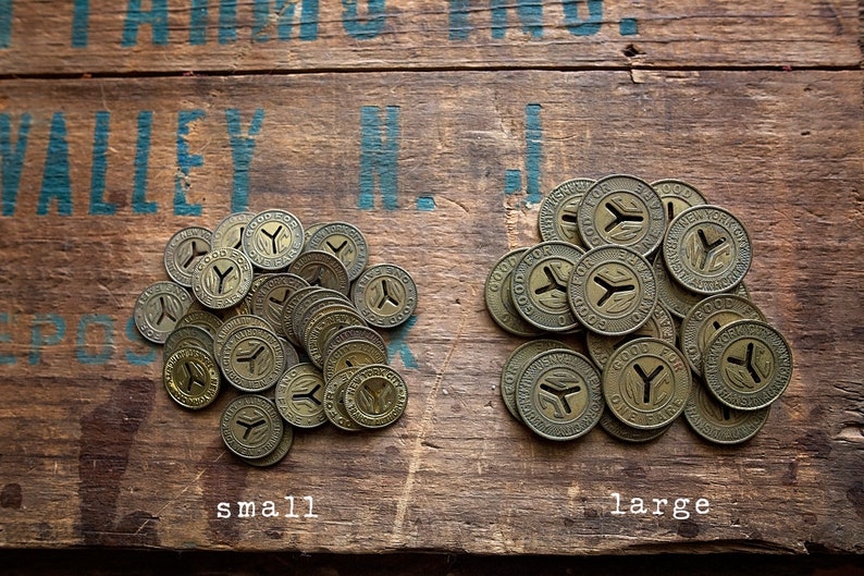 May include: Two piles of vintage brass tokens on a wooden surface. The tokens are round and have a Y-shaped design in the center. The text "GOOD FOR ONE FARE" is visible on the tokens. The pile on the left is labeled "small" and the pile on the right is labeled "large".