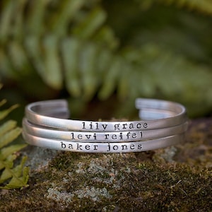 May include: Three silver cuff bracelets stacked on top of each other. Each bracelet is engraved with a name: "lily grace", "levi reifel", and "baker jones". The background is a blurred view of green foliage.