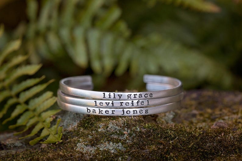 May include: Three silver cuff bracelets stacked on top of each other. Each bracelet is engraved with a name: "lily grace", "levi reifel", and "baker jones". The background is a blurred view of green foliage.