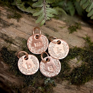 May include: Four copper-colored, round pendants with engraved letters and laurel leaf borders. Each pendant has a small loop for attaching to a chain or other item. The pendants are displayed on a mossy, wooden surface.
