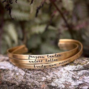 May include: Three gold-colored, engraved cuff bracelets stacked on a textured, mossy surface. Each bracelet is inscribed with a name: Grayson, Landon, Anders, Kellen, Brady, and Ava Maeve. The background is a blurred mix of green and brown.