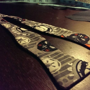 May include: Two gray fabric bow ties with a repeating pattern of black and white Star Wars themed graphics, including Darth Vader and the Imperial symbol. The ties are on a dark wooden surface.