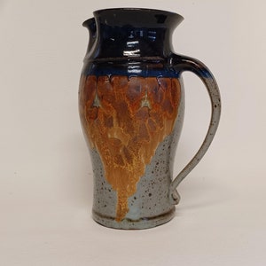 May include: A handmade ceramic pitcher with a dark blue rim and handle. The pitcher features a brown and orange design that drips down the center, with a speckled gray base. The pitcher has a curved handle and a spout.