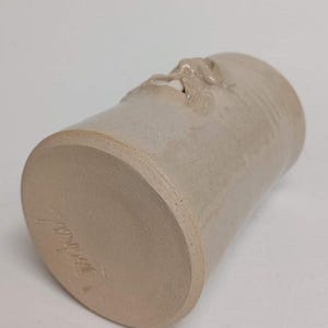 K&ouml;nnte beinhalten: Ein beigefarbener Keramikbecher mit einem geformten Gesichtsdesign in der N&auml;he der Oberseite. Der Boden des Bechers hat eine Signatur. Der Becher ist zylindrisch und hat eine leicht konische Form.
