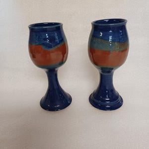 May include: Two handmade ceramic wine glasses. The glasses feature a dark blue rim, a band of red-orange, and a dark blue stem and base. The glasses are symmetrical and appear to be hand-painted.